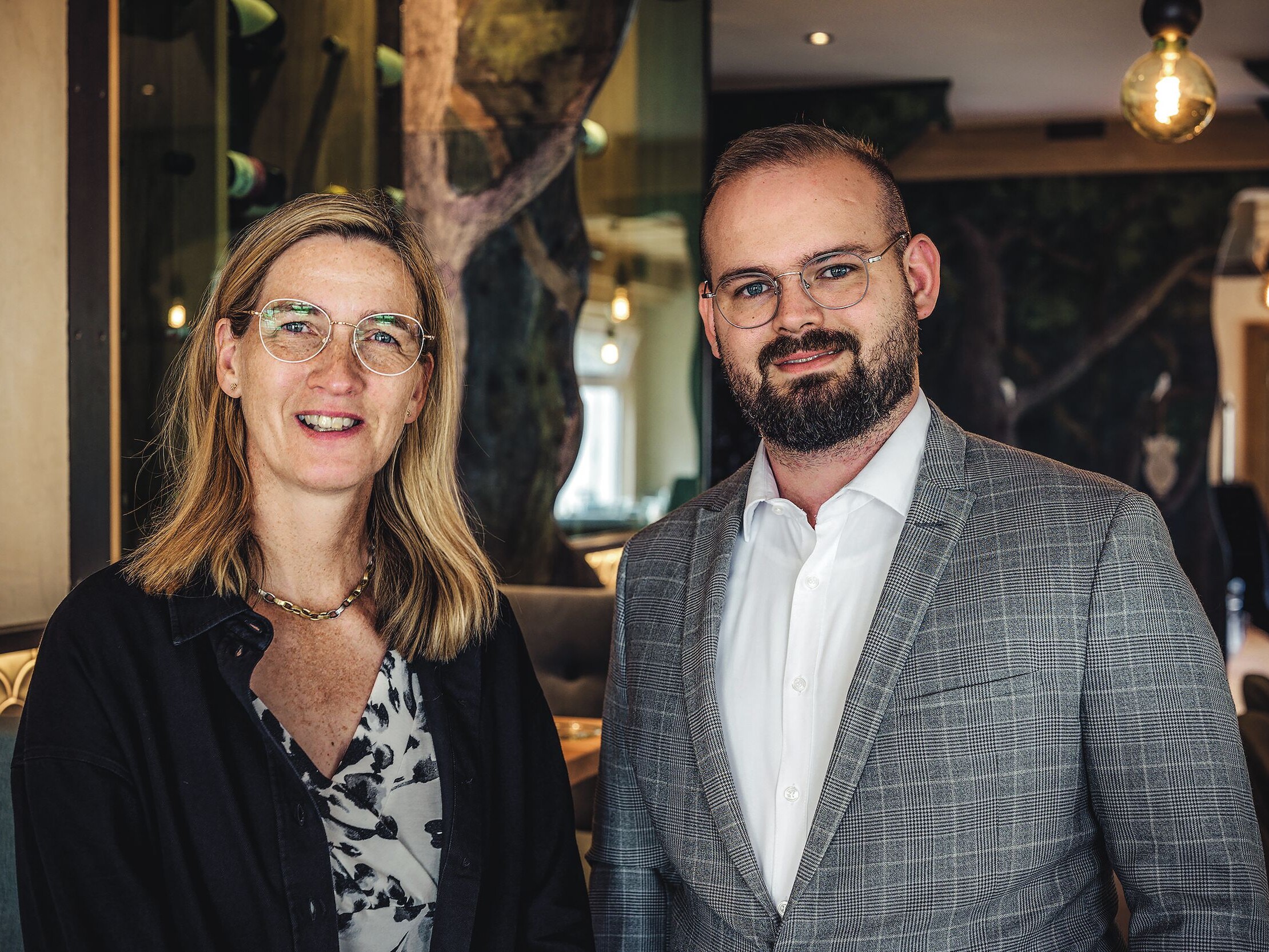 Sabine und Lukas Frankfurth Ein Mann und eine Frau stehen zusammen, beide lächeln, der Mann trägt einen Anzug und eine Brille.